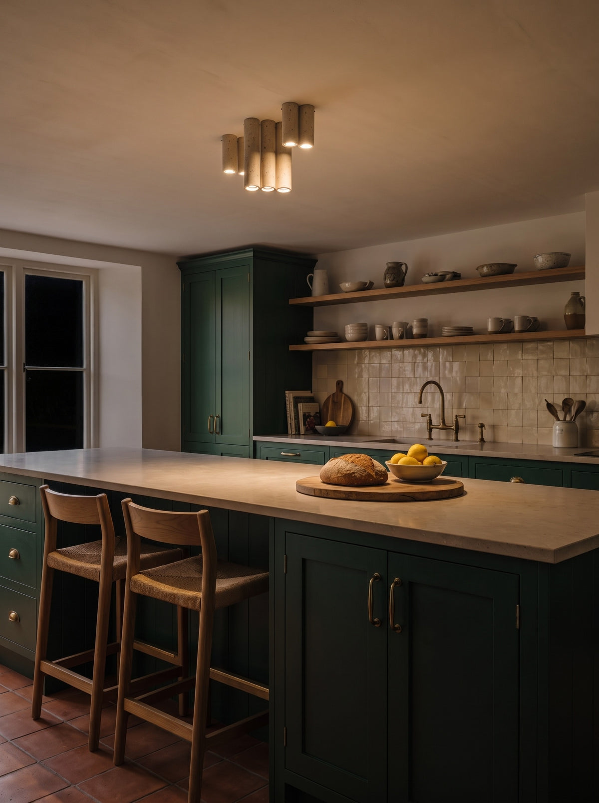 A cluster of seven travertine cylinder ceiling lights switched on above an island in a dark kitchen at night