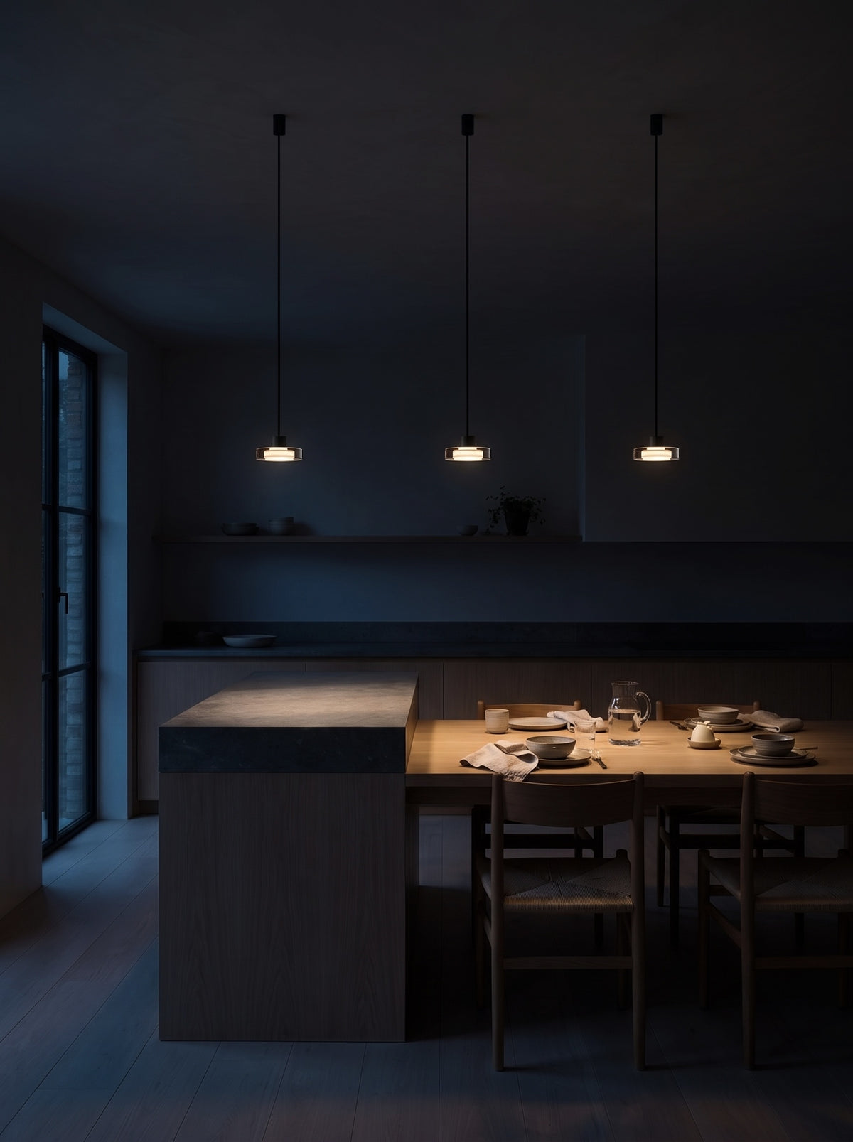 Three smoke grey glass pendant lights switched on above a kitchen island and dining table in a dark kitchen at night
