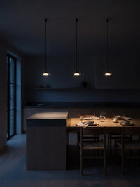 Three smoke grey glass pendant lights switched on above a kitchen island and dining table in a dark kitchen at night